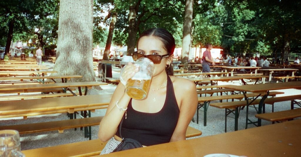 pexels-photo-30345991-30345991 A woman sipping beer at an outdoor beer garden during the day, surrounded by trees and empty benches.